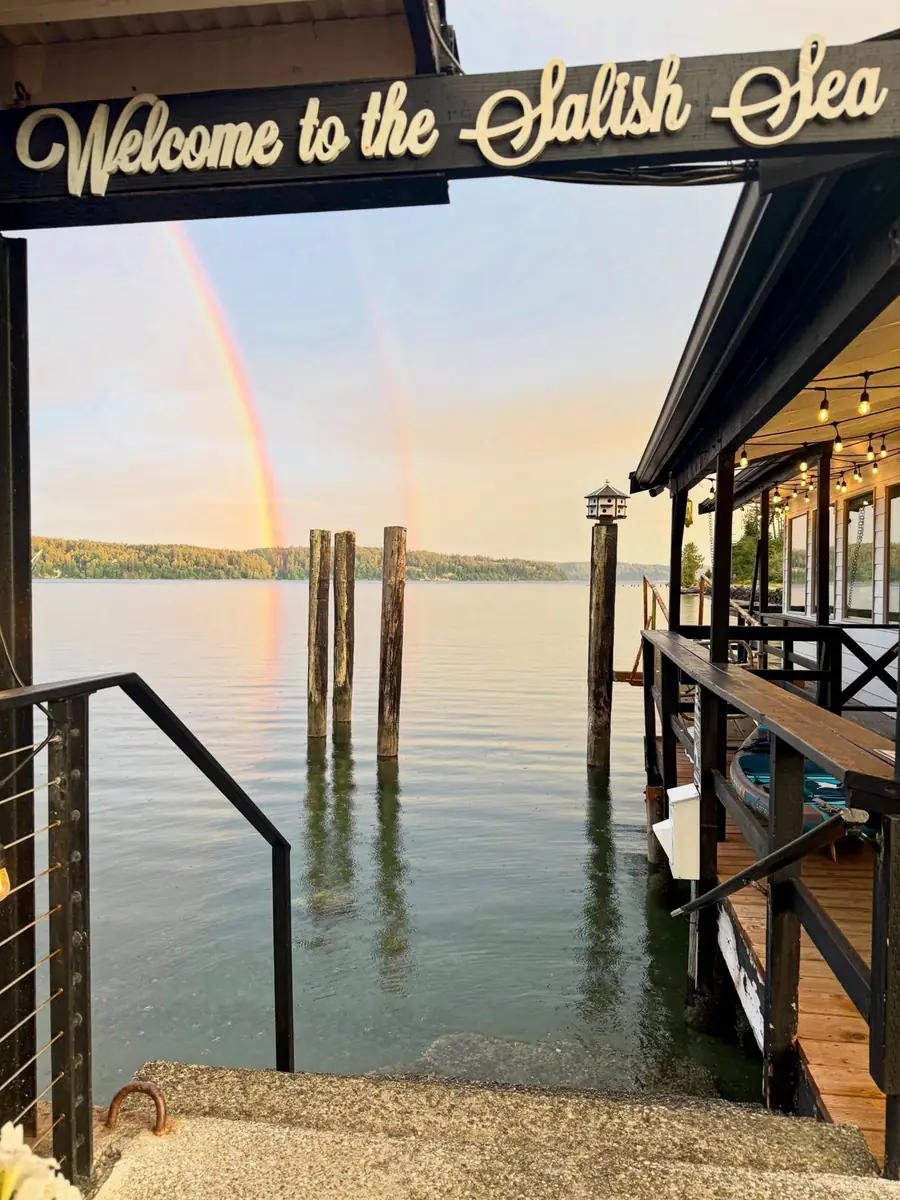 A double rainbow over Colvos Passage, framed by the market's Welcome to the Salish Sea sign and the old dock pilings