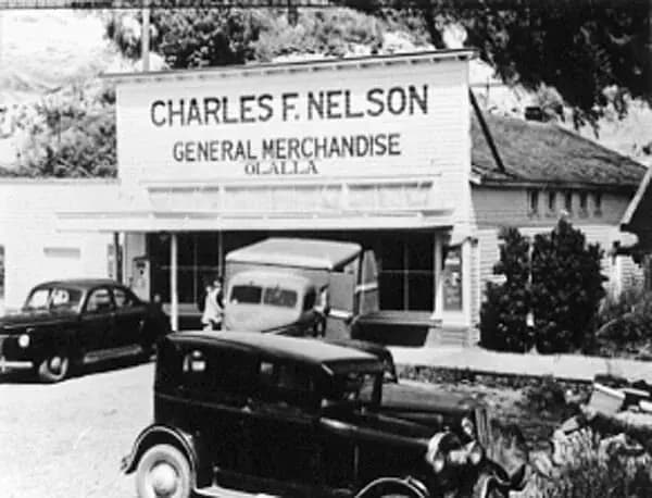 The white two-story Charles F. Nelson General Merchandise store with a delivery truck and cars out front
