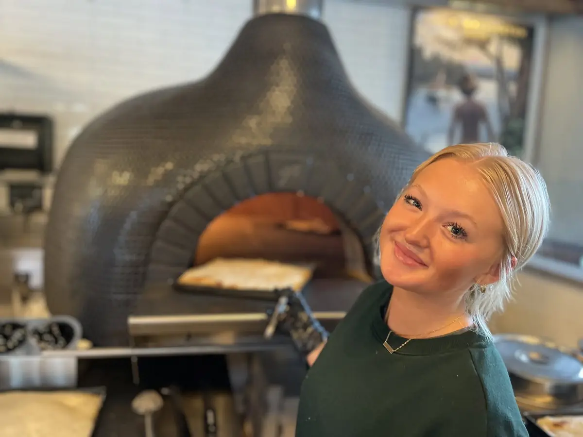 A staff member smiling in front of the tiled pizza oven as a pan pizza goes in