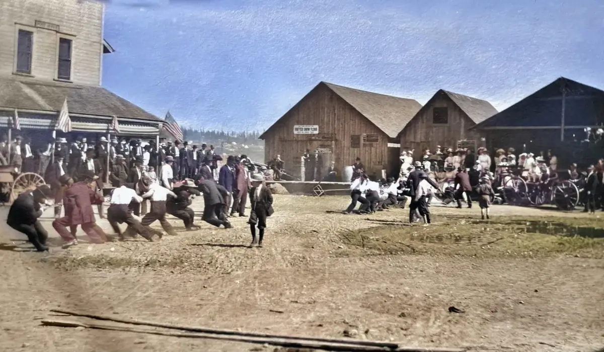 Colorized photo of a tug of war in the street, with flags on the store porch and a crowd watching